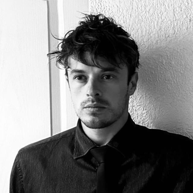 Black and white portrait of a man with tousled hair wearing a shirt and tie against a textured wall.