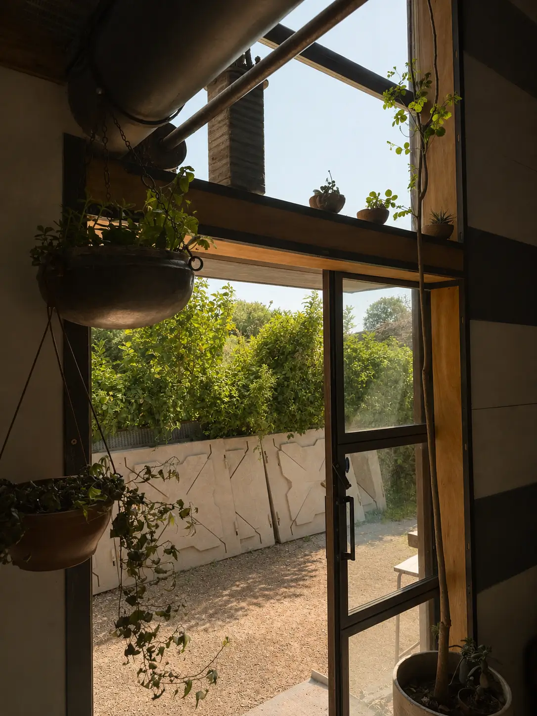 Open glass door framed with wood, showing sunny outdoor area with green bushes and hanging plants inside.