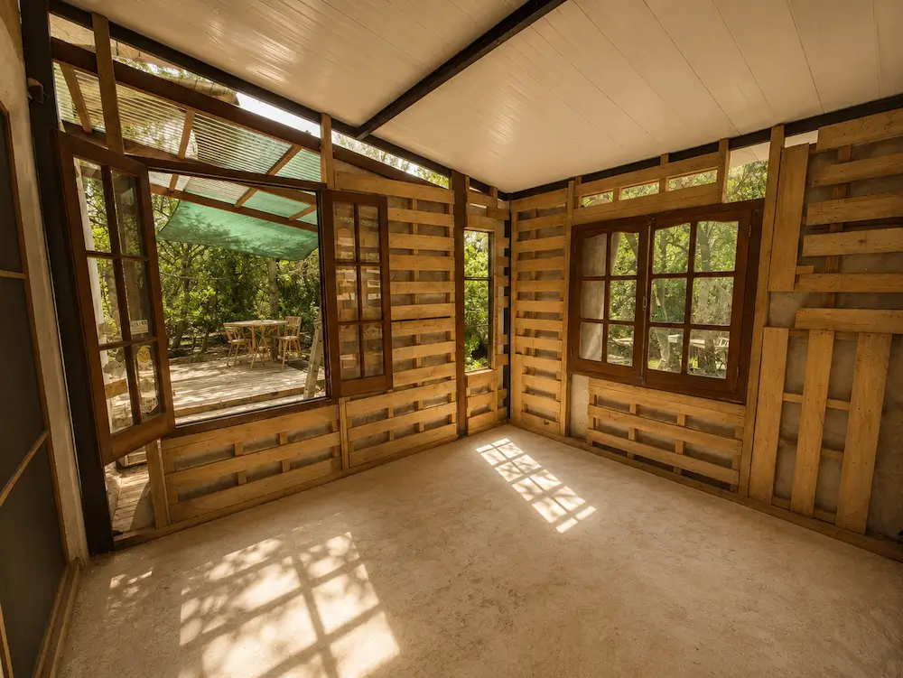 Empty room with unfinished wooden pallet walls, large windows, and sunlight casting shadows on the floor.