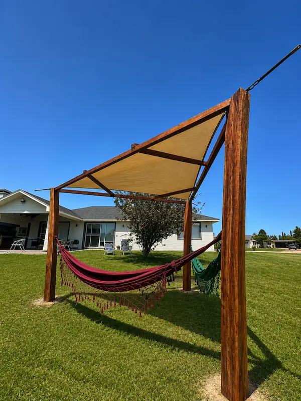 Wooden pergola with beige shade and two hammocks on green grass in a sunny yard.